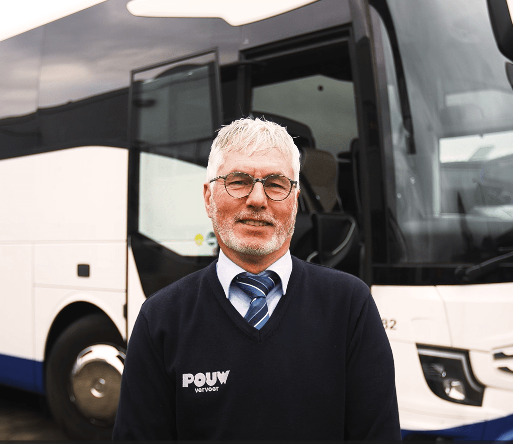 Marinus Rekers poseert voor zijn bus op het terrein van Pouw Vervoer. Hij draagt zijn uniform met trots en straalt werkplezier uit.